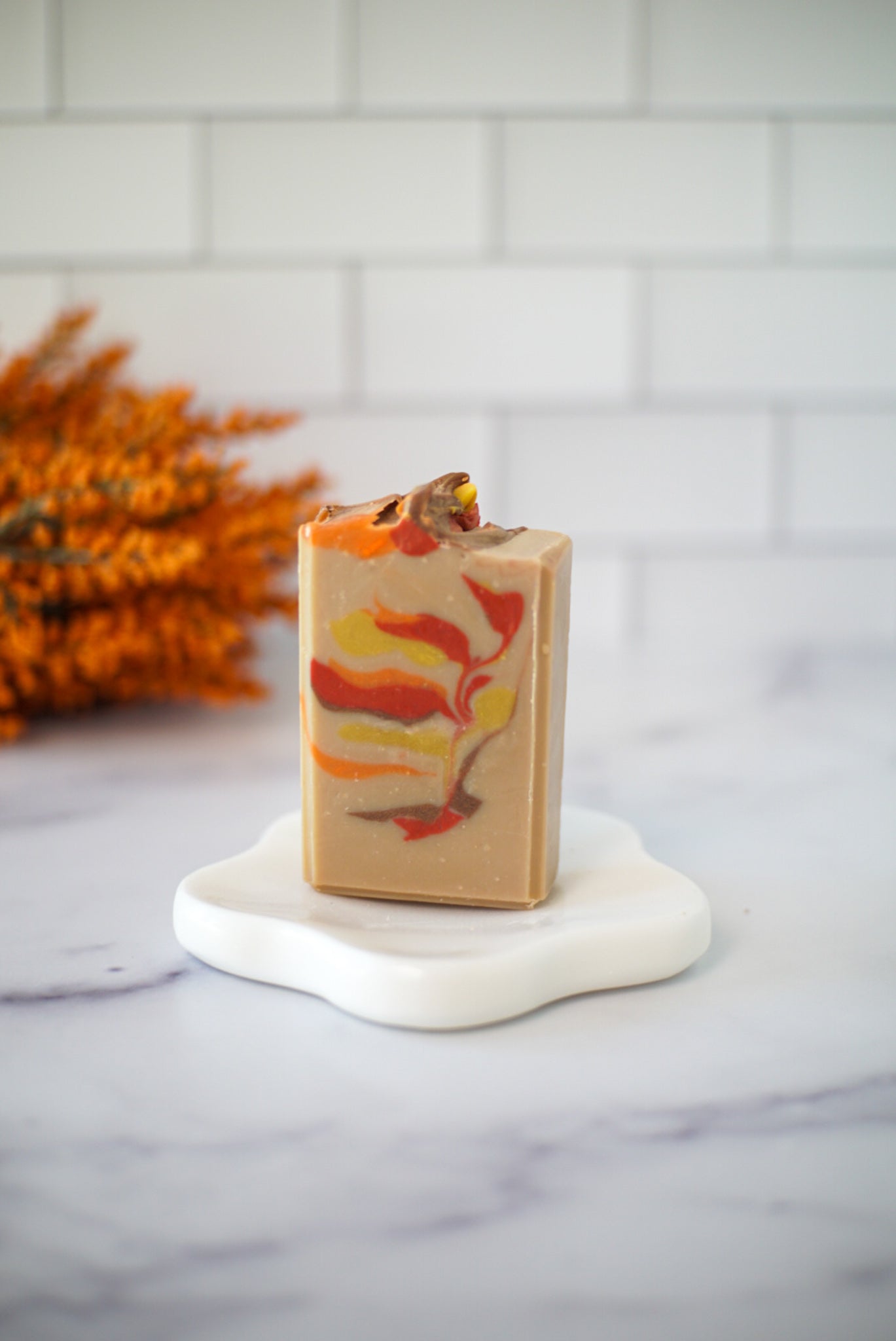 A rectangular bar of Flicker And Twinkle's New England Goat Milk Soap, adorned with vibrant orange, yellow, and red swirls, stands upright on a white ceramic flower-shaped dish. In the background, a blurry bunch of orange dried flowers contrasts against a white tiled wall. The soap rests on a smooth, light marble surface.
