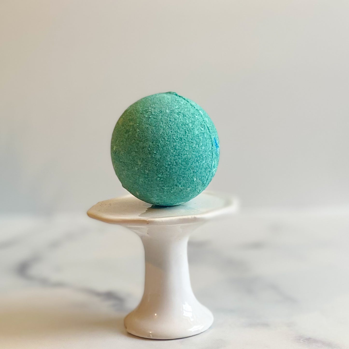 The Olive Branch Bath Bomb by Flicker And Twinkle, a blue-green sphere infused with fruity scents, sits atop a small white pedestal with a soft-focus marble background. The round bath bomb and the minimalist pedestal create a simple yet elegant composition.