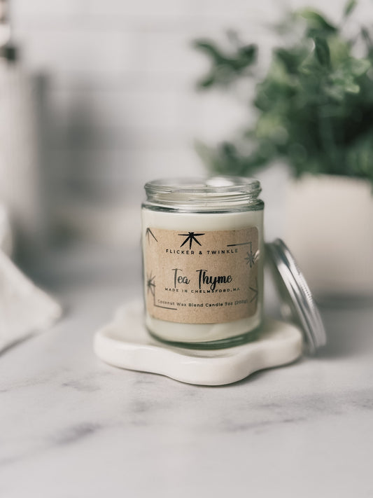 Tea Thyme Coconut Wax Candle with lid off sits on a coaster on a marble surface.