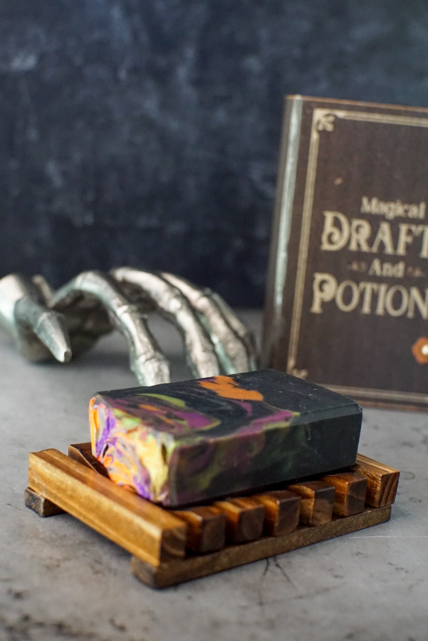 A decorative scene showcasing the "That Witch Goat Milk Soap" by Flicker And Twinkle, featuring vibrant swirls of colors and placed on a wooden soap dish. In the background, a prop spell book titled "Magical Drafts And Potions" and a skeletal hand decoration capture the essence of spooky season soap on a gray surface.