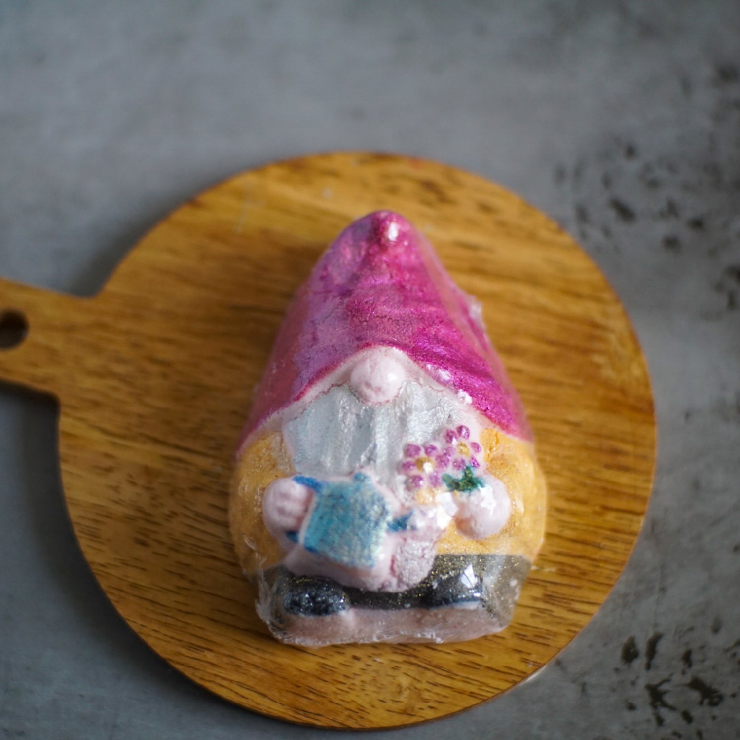 A small gnome figurine with a pink hat sits on a round wooden board, holding a book and flowers. The background is a neutral gray surface.