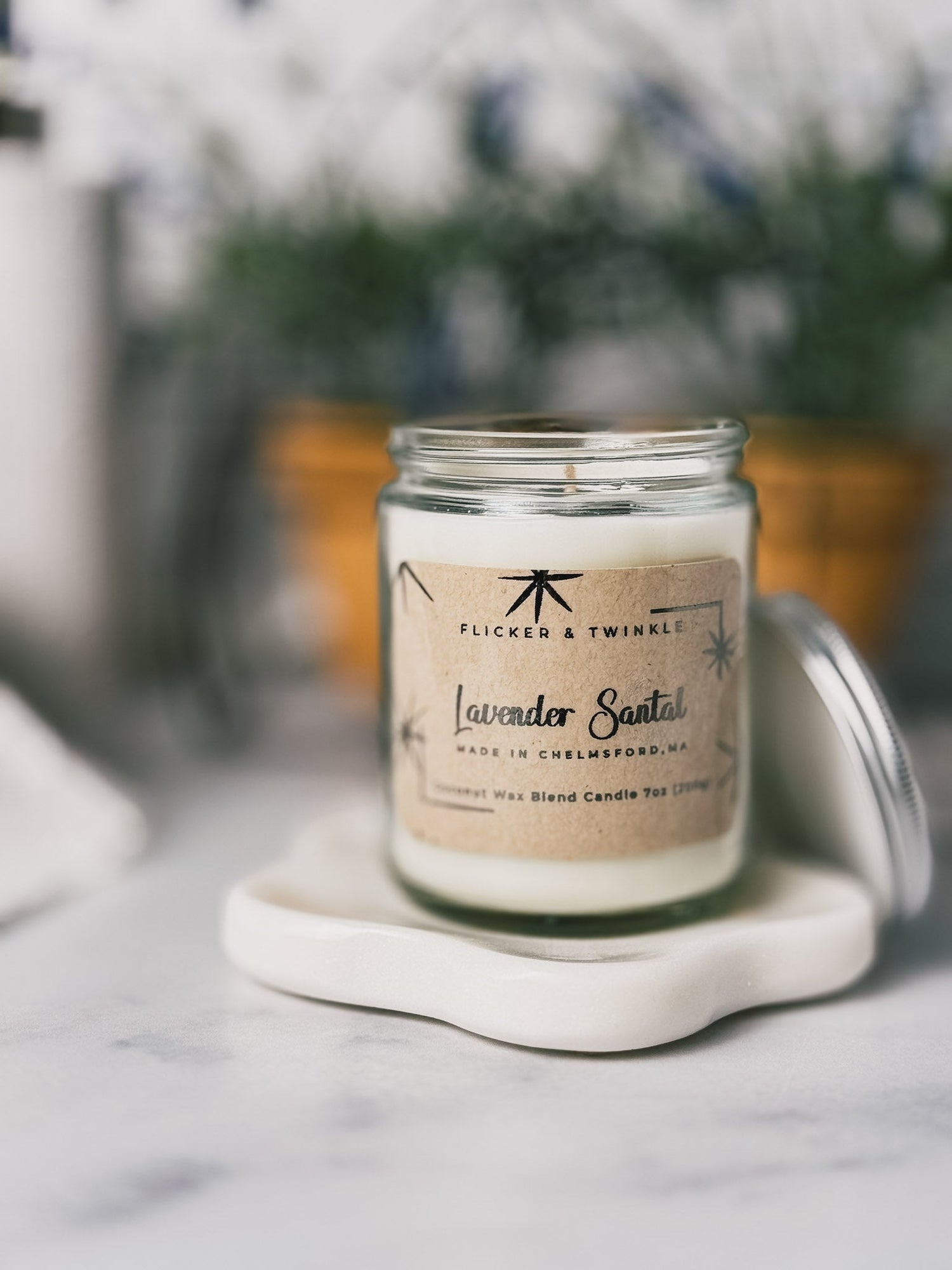 A glass jar candle labeled "Lavender Santal" on a white coaster. The background is softly blurred, with a hint of greenery and a pot. The scene feels calm and inviting.