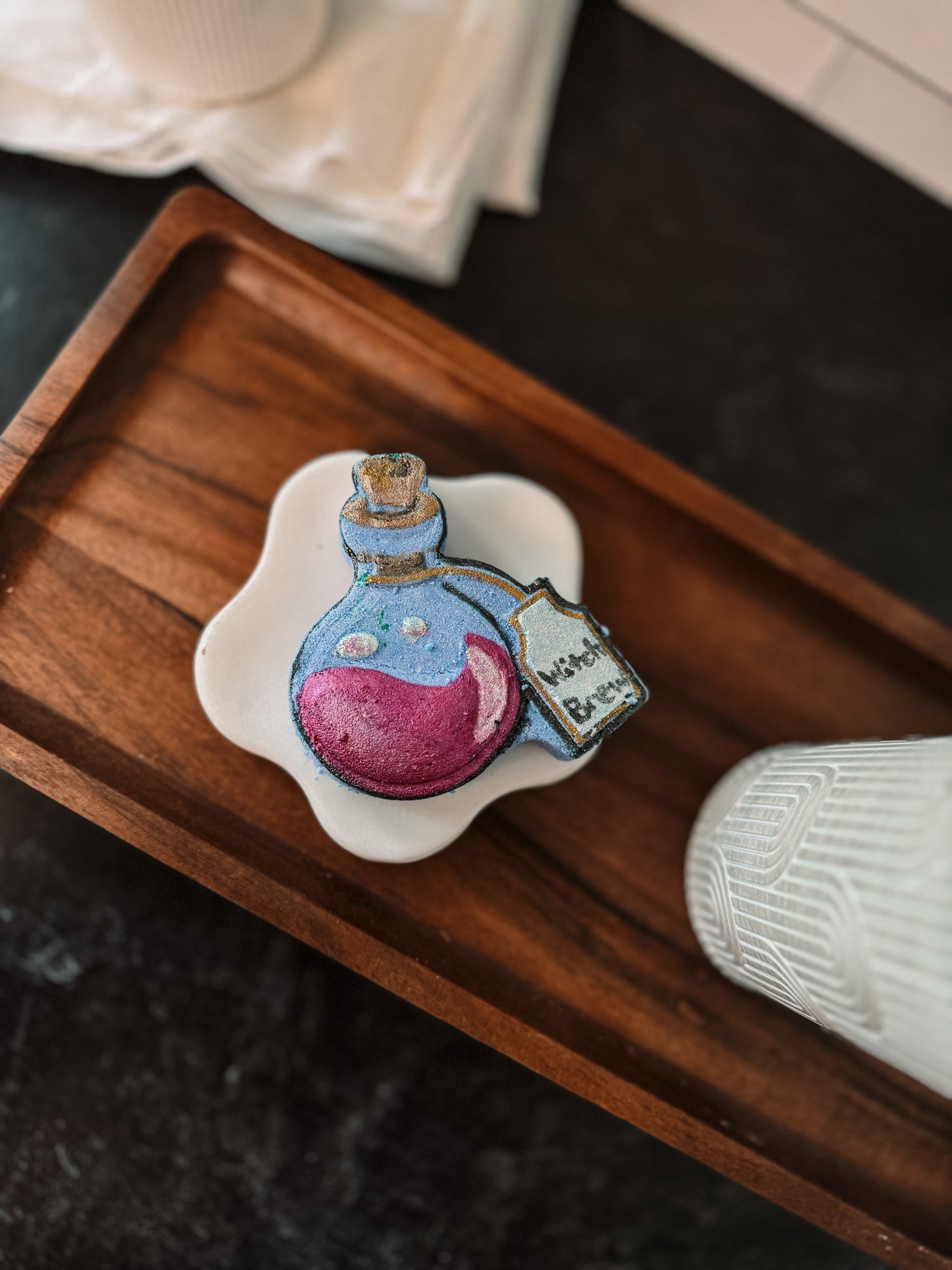 A wooden tray holds a small, decorative item resembling a potion bottle with a cork stopper and purple liquid inside. It has a tag reading "love potion." A part of a white cup is visible next to it.