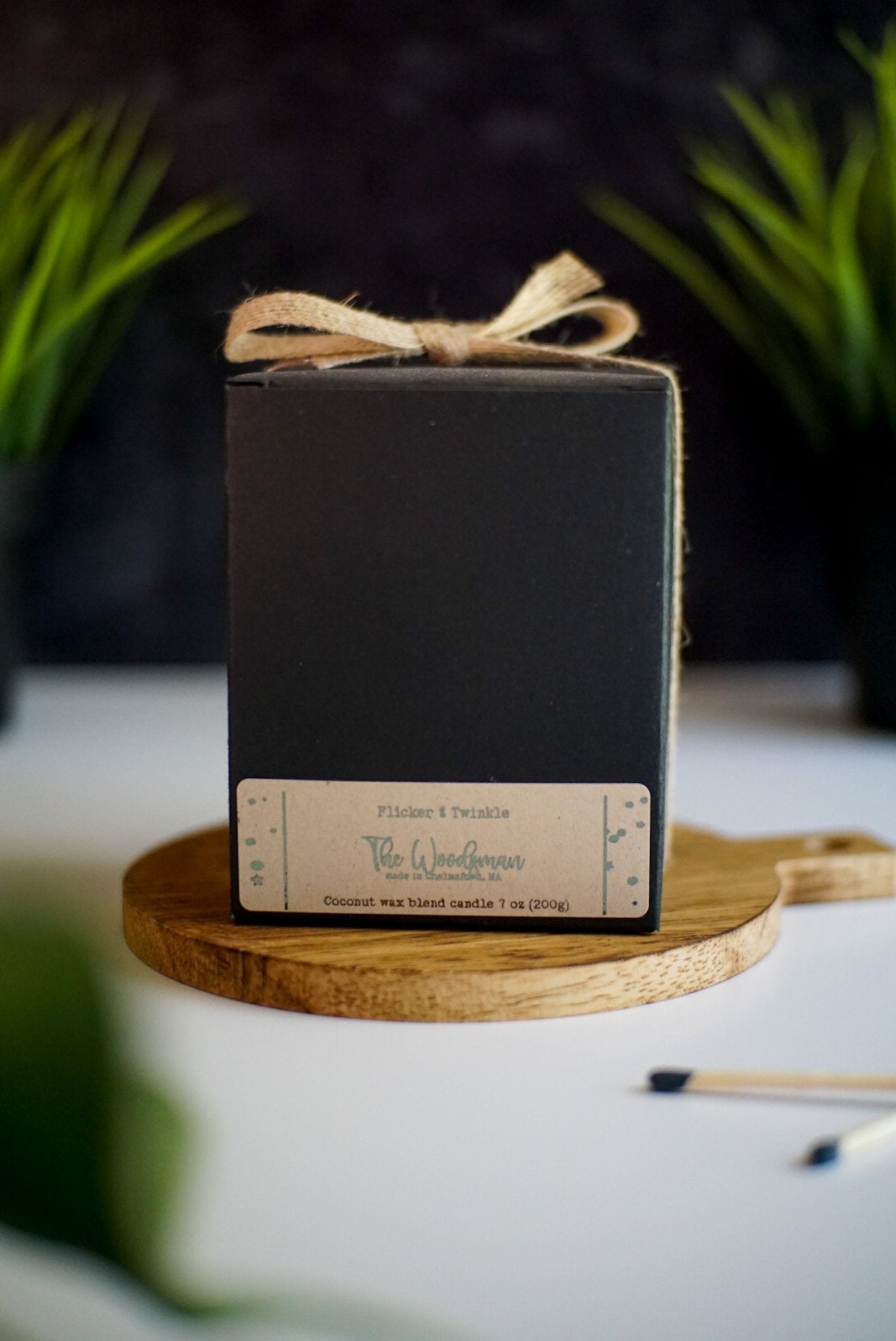 A neatly wrapped black box with a bow, labeled "Flicker + Twinkle The Woodsman," sitting on a wooden platform. Surrounded by small plants and matchsticks, it exudes a rustic, natural vibe.
