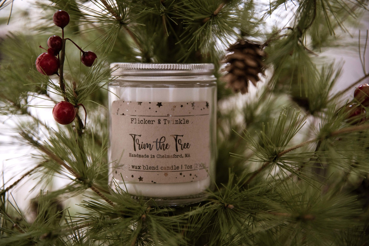 Close-up of a "Trim the Tree" candle by Flicker & Twinkle, nestled among pine branches with red berries and a pinecone. The candle is handmade in Chelmsford, MA and weighs 7 ounces.