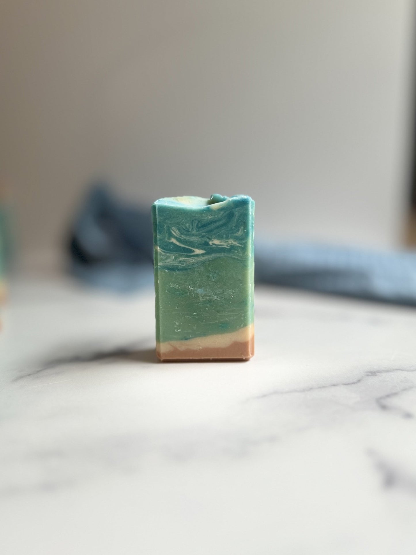 The Lagoon Goat Milk Soap by Flicker And Twinkle, a rectangular bar of nourishing shea butter, stands upright on a light, smooth surface. The soap features beautiful layers of teal, green, and beige marbling. In the background, there's a subtle blur with hints of a blue cloth and a gradient from light to dark.