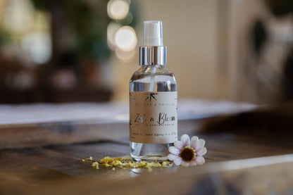 A glass bottle of Flicker And Twinkle's Zest and Bloom Room and Linen Spray, featuring phthalate-free ingredients and refreshing citrus scents, sits elegantly on a wooden table. The bottle has a silver spray nozzle and is adorned with small yellow petals and a single daisy-like flower at its base. The background is softly blurred.