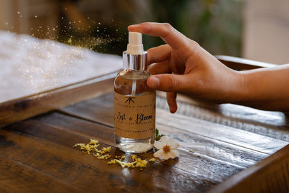 A hand is shown spraying a bottle labeled "Flicker And Twinkle Zest and Bloom Room and Linen Spray," surrounded by small white flowers and yellow petals on a wooden tray. The mist of droplets, filled with citrus scents and phthalate-free ingredients, disperses into the softly blurred background.