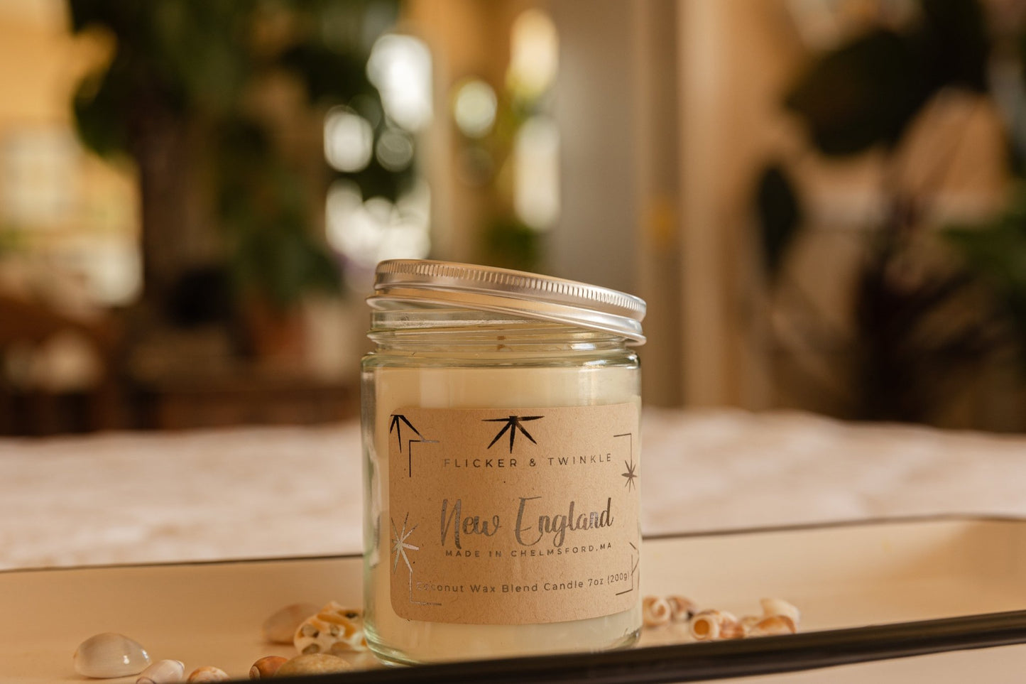 A glass jar candle labeled "Flicker And Twinkle" sits on a surface surrounded by small seashells. The label reads "New England Coconut Wax Candle" and "Coconut Wax Blend Candle 7oz (198g)." The background appears blurred with indoor plants and soft lighting.