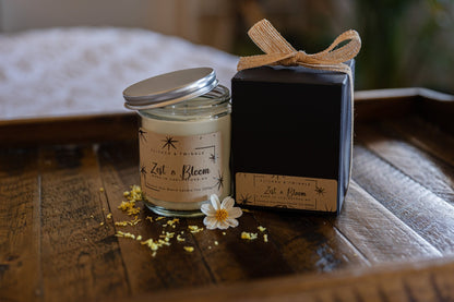 A lit Zest and Bloom Coconut Wax Candle by Flicker And Twinkle, featuring a refreshing lemon mint lilac scent and housed in a glass jar with the "Zest & Bloom" label, sits on a wooden tray next to a black gift box adorned with a beige ribbon. A small white flower and scattered petals are placed near the clean-burning candle.