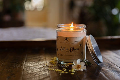 A lit Zest and Bloom Coconut Wax Candle by Flicker And Twinkle, housed in a glass jar, sits on a wooden surface. Its metal lid leans against the jar. Nearby, small yellow flowers and a white daisy add to the ambiance. The background features softly blurred green plants with light streaming in from a window.