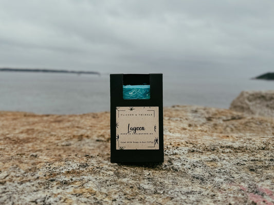 A black box containing a bar of "Lagoon Goat Milk Soap" by Flicker And Twinkle rests on a rock near the sea. The minimalist design showcases stars and text on the label. Infused with nourishing shea butter, it sits against a backdrop of cloudy sky and distant shoreline, embodying serene sea salt vibes.