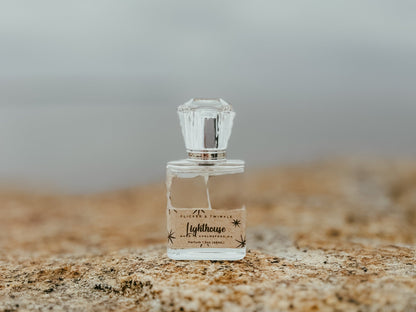 A clear glass perfume bottle labeled "Lighthouse Parfum" from Flicker And Twinkle, adorned with a star design, rests on a textured rock surface, exuding the potent essence of Parfum. The bottle features a faceted, diamond-shaped cap. The background is softly blurred, evoking the scent of ocean air and a serene coastal atmosphere.