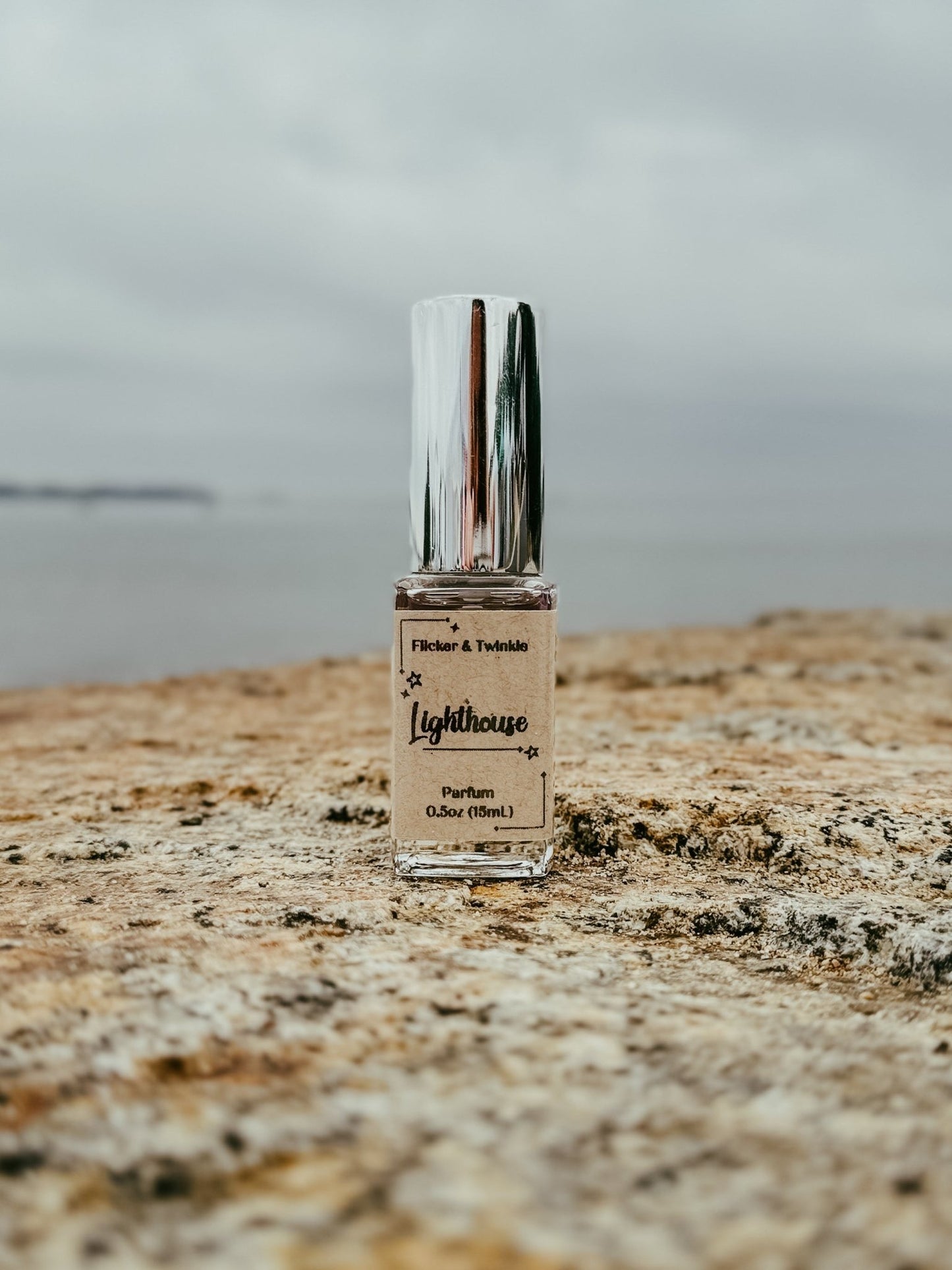 A bottle of "Lighthouse Parfum" from Flicker And Twinkle is placed on a rough stone surface, set against an overcast sky and distant shoreline. Capturing the essence of ocean air, the bottle features a reflective silver cap and holds 0.5 ounces (15 ml) of parfum strength fragrance.