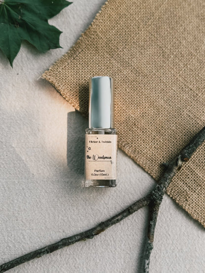 A small bottle of The Woodsman Parfum by Flicker And Twinkle, adorned with a silver cap, rests on a piece of burlap. A branch and part of a green leaf in the background complete the earthy woods setting, exuding rustic and natural charm.