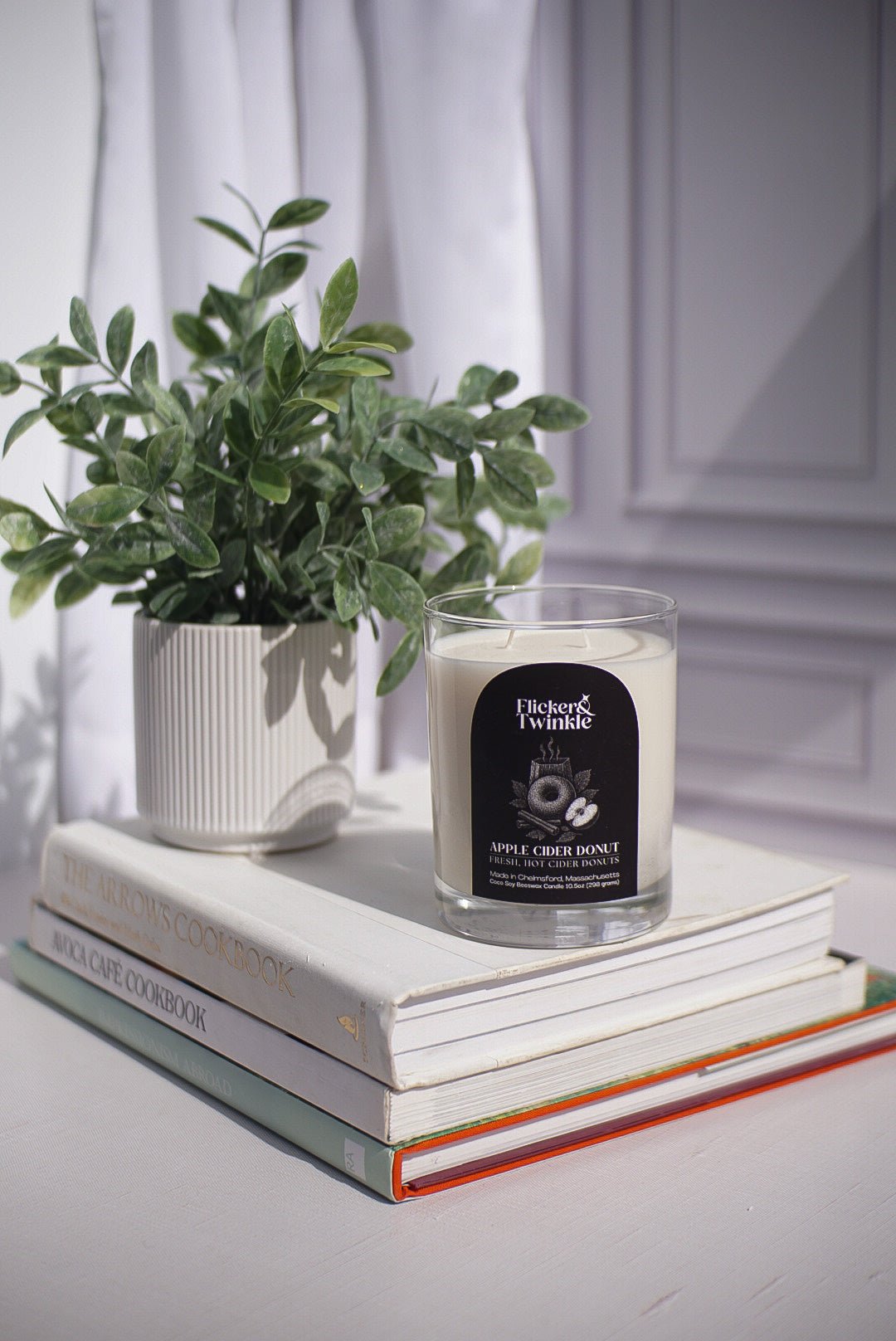 An Apple Cider Donut Candle sits atop three cookbooks on a white table with a potted plant.