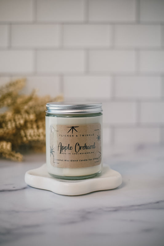 A handcrafted jarred candle labeled "Apple Orchard" sits on a marble surface with a blurred autumnal decoration in the background, promising a clean-burning and long-lasting glow.