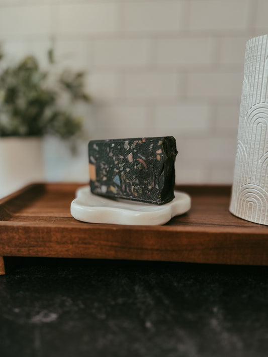 A bar of black and white marbled goats milk soap on a ceramic dish, placed on a wooden tray, with a blurred plant and textured container in the background.