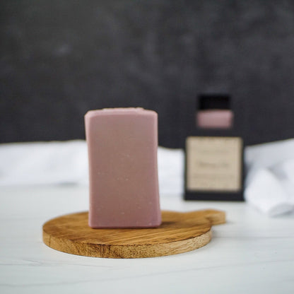 A bar of light purple handmade soap stands upright on a small, round wooden tray. In the blurred background, a matching soap box is partially visible, along with a white cloth on a white surface and a dark wall. The scene has a minimalistic and clean aesthetic, evoking the purity of goat's milk soap.