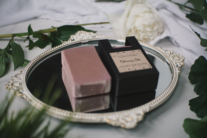 A rectangular, pink soap bar and its black packaging labeled "Blooming Lilac" are placed on an ornate, silver tray. The tray is surrounded by green leaves and a white flower on a white fabric background. This is a luxury Goat Milk Soap