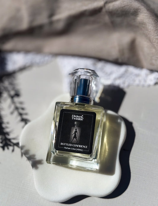 A square glass perfume bottle labeled 'Bottled Confidences' with a black label and silver cap, resting on a white ceramic dish against a neutral background. Orange Blossom and Mandarin elegant Parfum. 