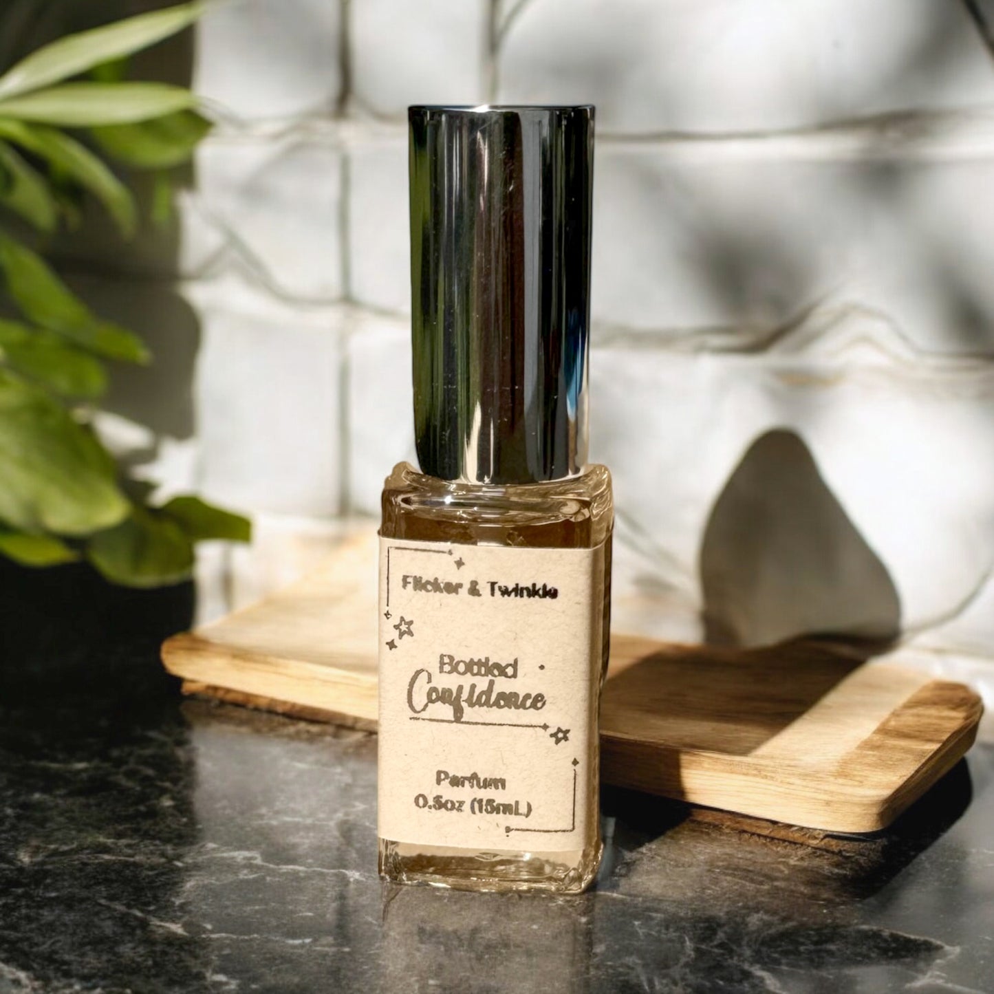 A small bottle of Bottled Confidence Perfume by Flicker And Twinkle is placed on a wooden surface. The transparent bottle with a shiny silver cap is illuminated by natural light, with green foliage and white tile in the background, hinting at notes of Citrus Energy and Orange Blossoms.