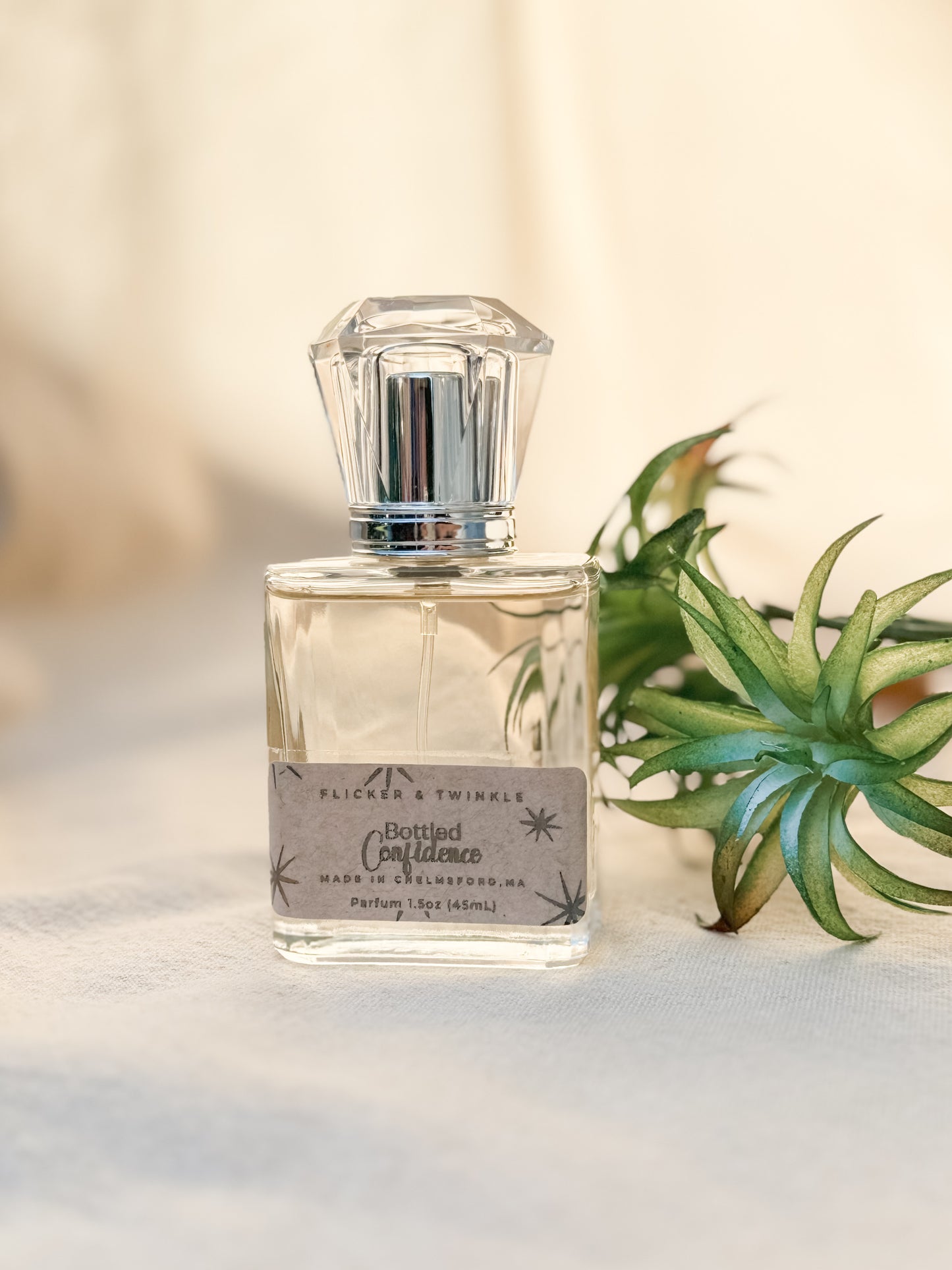 A clear glass perfume bottle with a faceted cap, labeled "Bottled Confidence Perfume" from Flicker And Twinkle, is partially filled with a light-colored liquid. It rests on a soft, white, textured surface next to a small green succulent. The fragrance promises an invigorating burst of Citrus Energy intertwined with delicate Orange Blossoms.