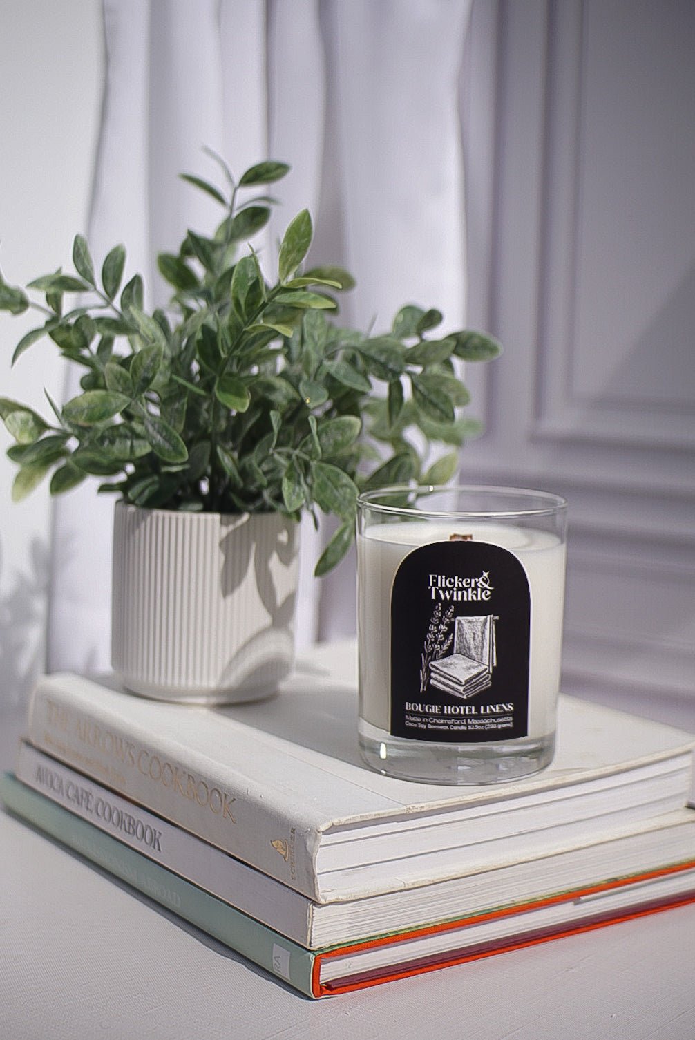 A Bougie Hotel Linens Candle - Coconut Soy Beeswax sits atop cookbooks by a white curtain.