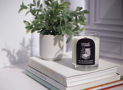 A Bougie Hotel Linens Candle - Coconut Soy Beeswax sits on books beside a potted plant.