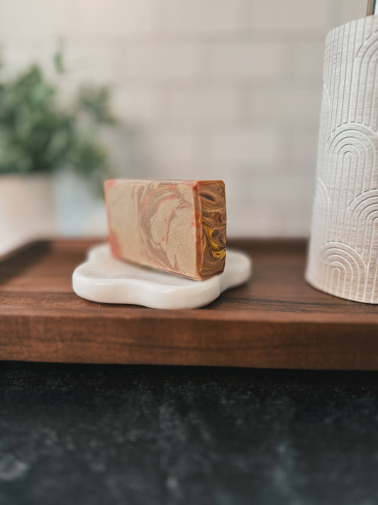 A rectangular bar of Flicker And Twinkle's Caramel Apple Goat Milk Soap, featuring swirls of beige, brown, and gold, is placed on a white soap dish shaped like a cloud. The soap dish rests on a wooden surface. Background elements include a plant and a textured dispenser.