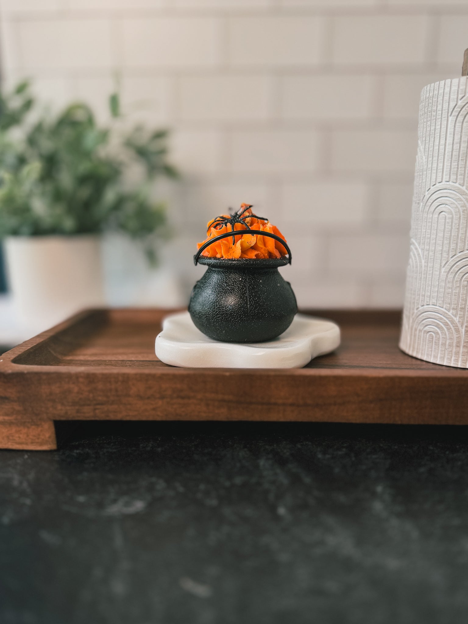 A Flicker And Twinkle Bath Bomb Cauldron, small and black, is filled with bright orange piped bubble bath and placed on a white ceramic stand atop a wooden tray. The background features a blurred plant and a textured white cylindrical object, evoking hints of fruity magic. This enchanting scene is set on a dark countertop with a white tiled wall in the background. Perfect for a Halloween gift