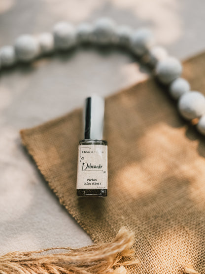 A small bottle of Debonair Parfum from Flicker And Twinkle rests on a piece of burlap fabric. The background features a blurred strand of round white beads, and soft natural light creates a serene, rustic atmosphere. As part of a luxury summer fragrance collection, the bottle boasts a silvercap and an elegant label design.