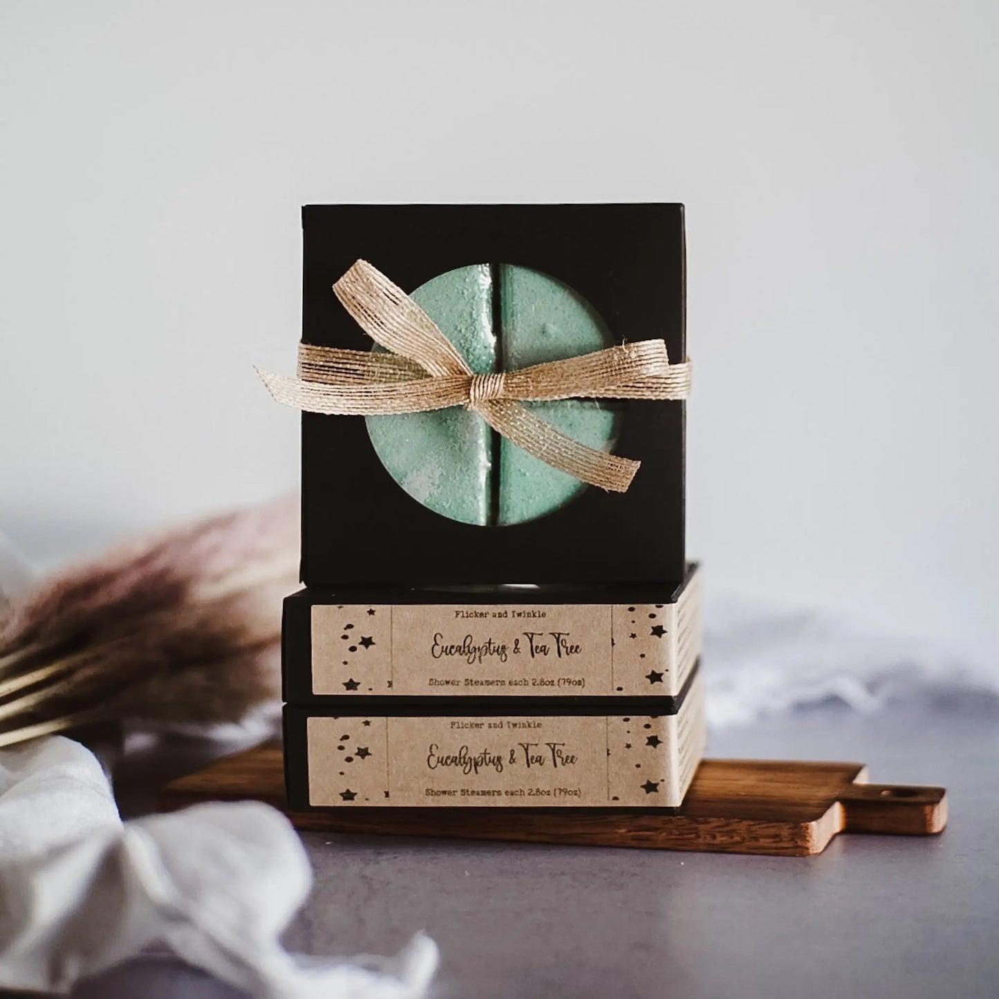 A rustic arrangement featuring two brown boxes labeled "Flicker and Twinkle" with "Everything's No. One" butter slammers with lotus. They are topped with a black box containing a green item, tied with a textured beige ribbon. Shower Steamers and pampas grass add to the spa-style escape ambiance.