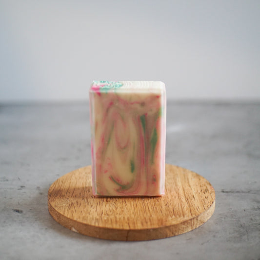 A handmade, rectangular bar of Flicker And Twinkle's Fig and Pink Pepper Vegan Soap with a marbled design in pink, green, and beige hues sits on a round wooden platform against a grey background, exuding a luxurious scent.