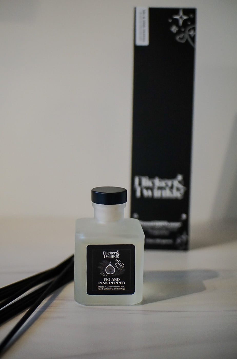 Fig and Pink Pepper scented Reed diffuser bottle with black cap and label on a light surface, with a blurred black box in the background.