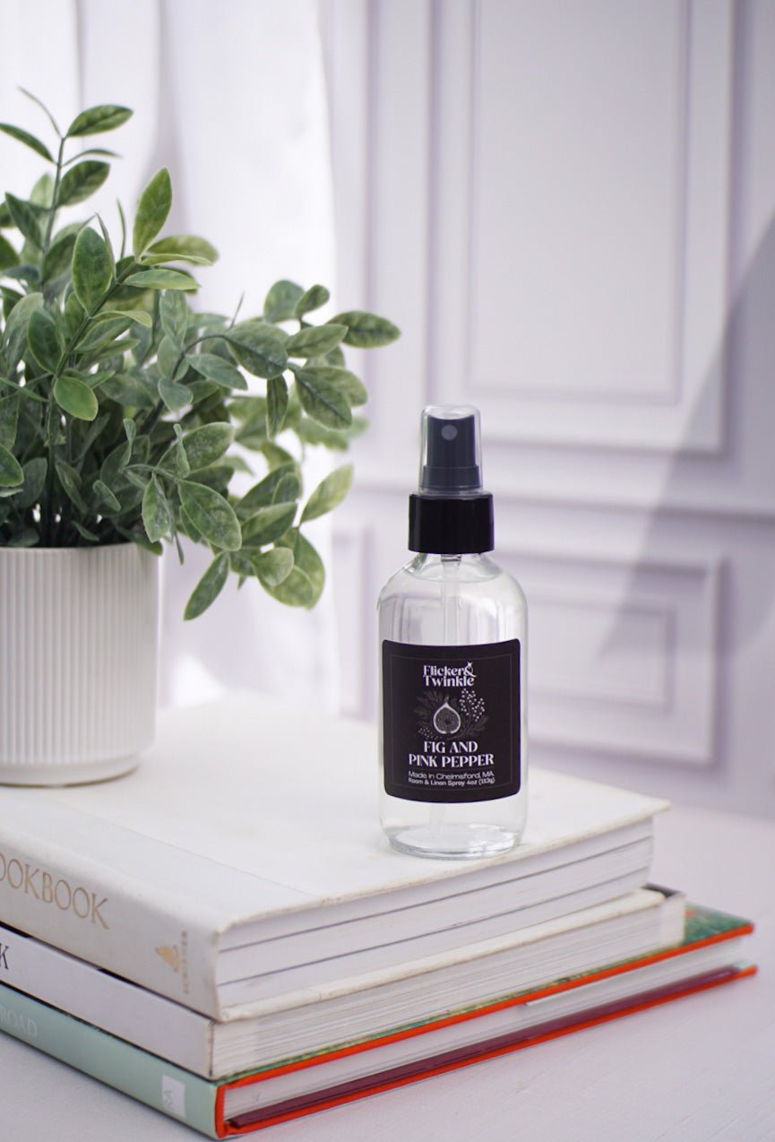 Fig and Pink Peppercorn Room and Linen Spray on books beside a plant in a bright room.