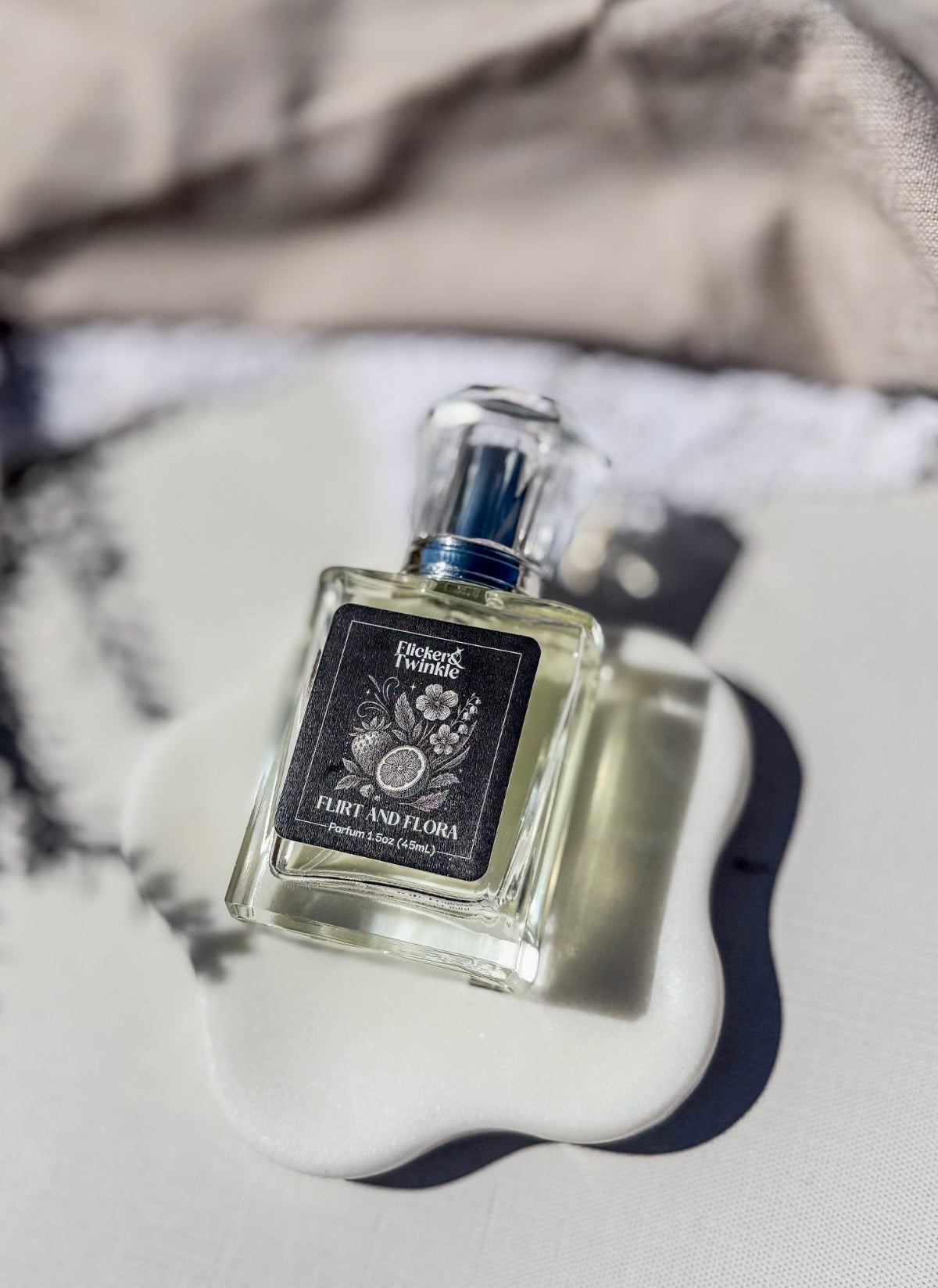 A bottle of perfume with a black label reading 'Flirt and Flora" sits on a white ceramic dish against a neutral, softly blurred background.