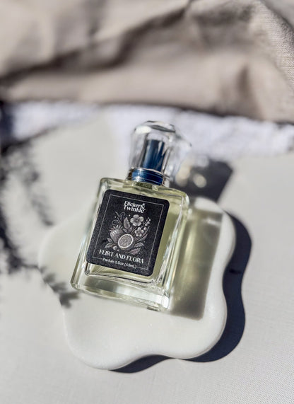 A bottle of perfume with a black label reading 'Flirt and Flora" sits on a white ceramic dish against a neutral, softly blurred background.