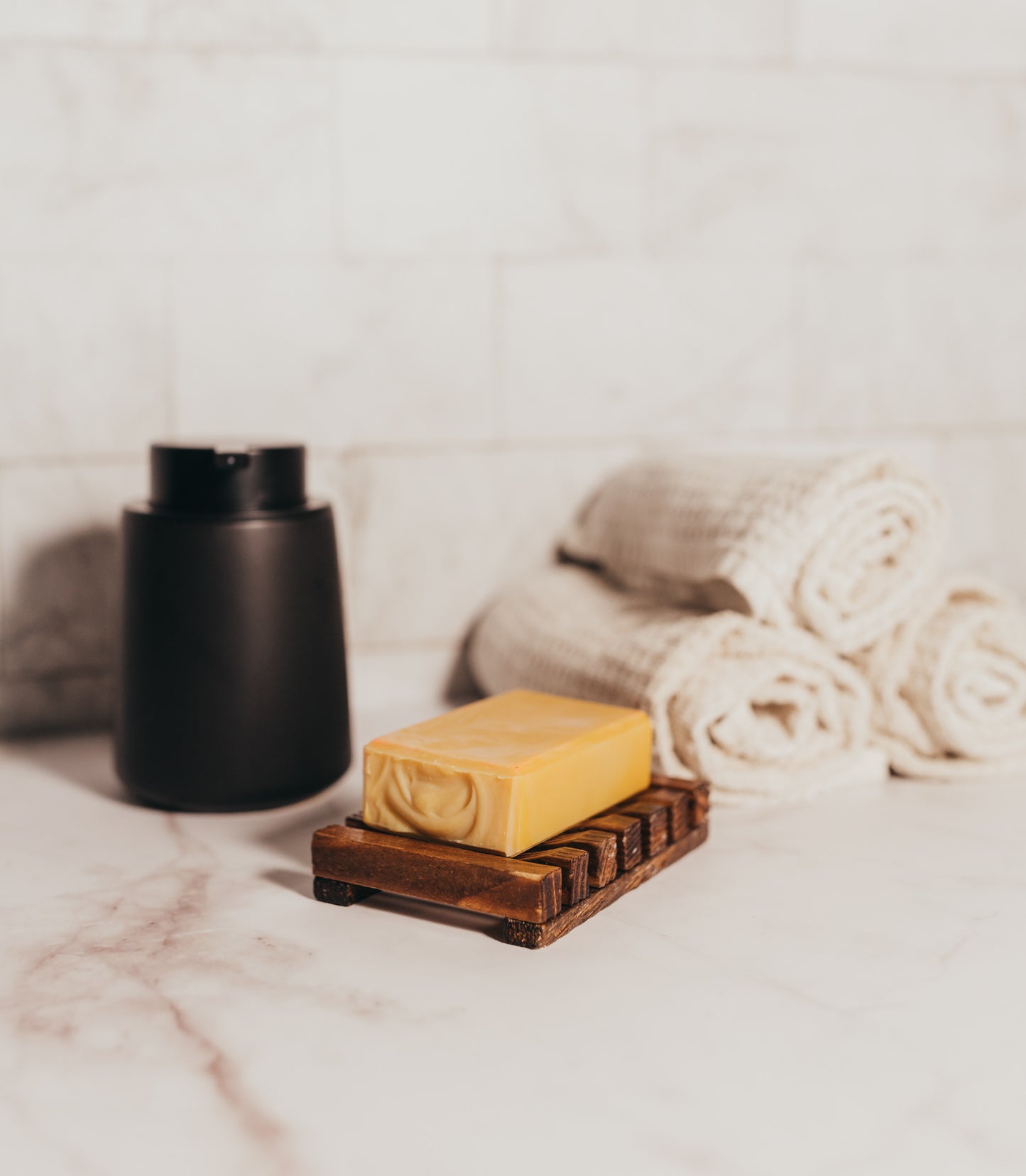 A wooden soap dish holds a bar of "From Sorrento with Love" goat's milk soap by Flicker And Twinkle on a marble countertop. Behind the soap dish, there are neatly rolled, textured white towels. To the left, there is a matte black cylindrical container. The background features a white tiled wall.