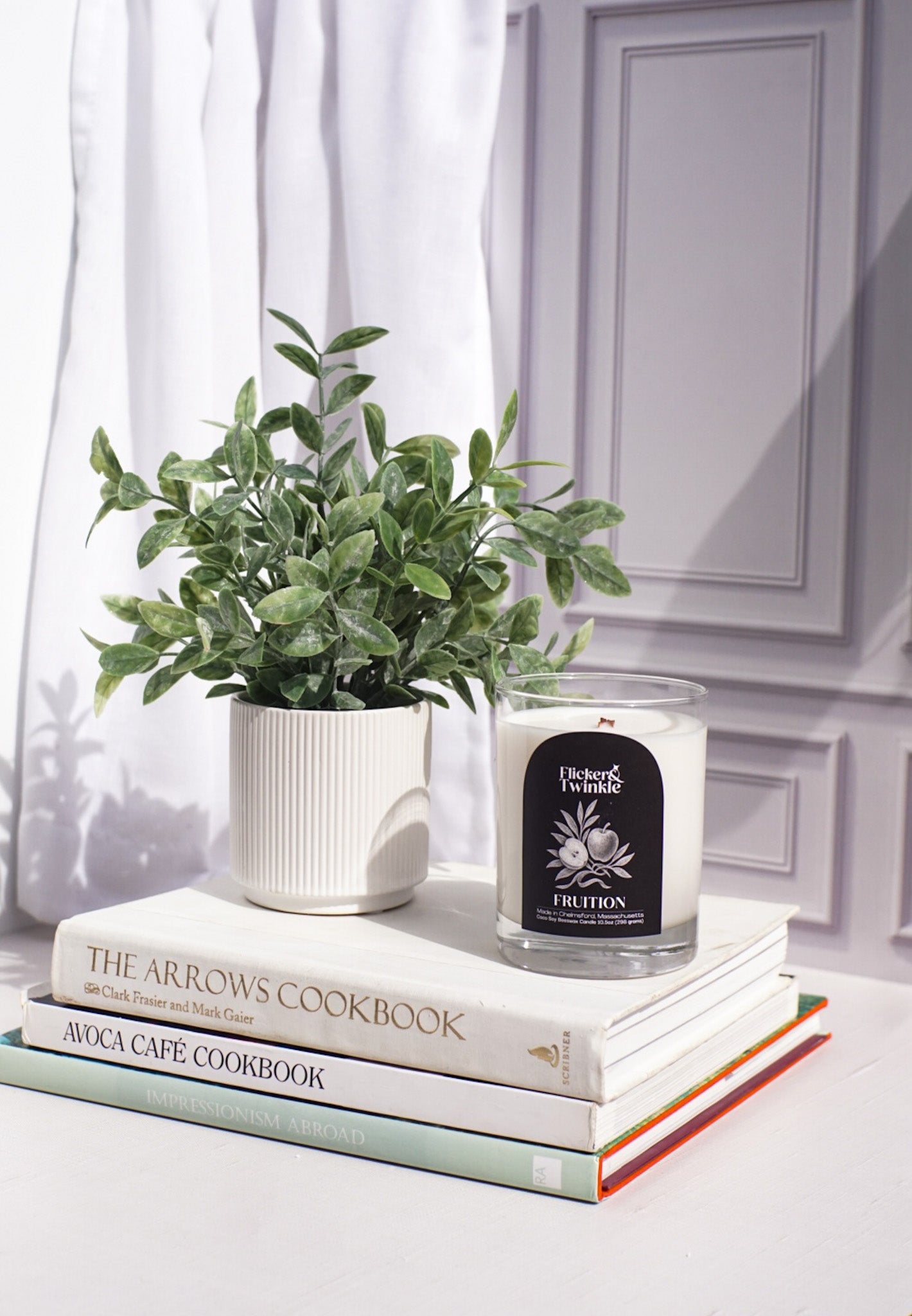 A Fruition Candle – Coconut Soy Beeswax and a potted plant on cookbooks by a window.