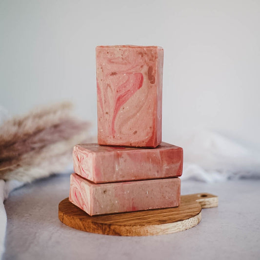 Three rectangular bars of Flicker And Twinkle's Girls Trip Goats Milk Soap, featuring a pink marbled design, are stacked on a small, round wooden board. The soap bars have a natural, handmade appearance and are set against a neutral background with soft fabric and dried plant elements, evoking an opulent spa experience.