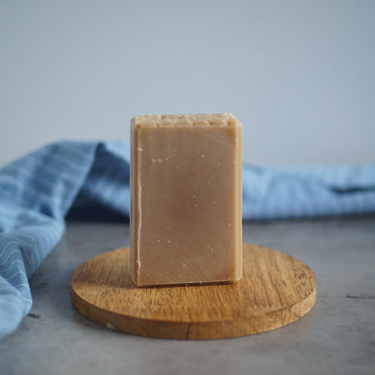 A luxurious bar of Flicker And Twinkle's Honeyed Oats Goat Milk Soap stands upright on a round wooden dish. The beige soap, enriched with nourishing goat's milk, boasts a smooth, slightly textured surface. In the background, a light blue cloth with a striped pattern is draped over a gray surface. The overall scene is minimalist and clean.