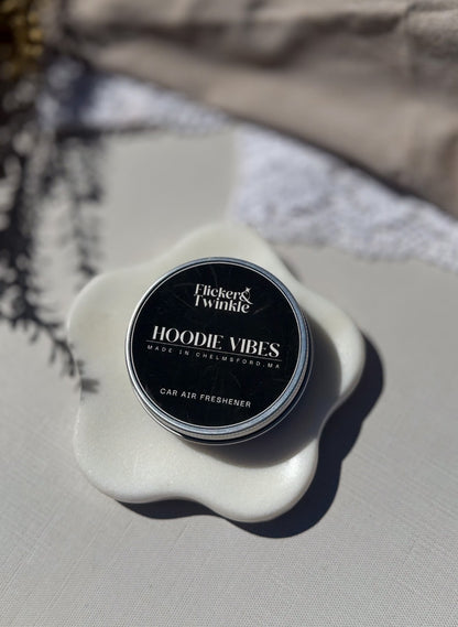A round tin of Flicker & Twinkle Hoodie Vibes car air freshener sits on a white ceramic flower-shaped dish. The product has a black label with white text, set against a neutral beige fabric background with soft shadows.