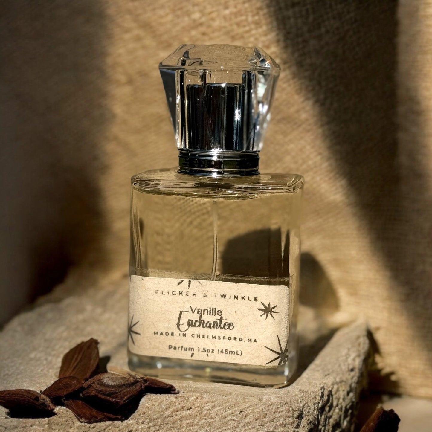 A square glass perfume bottle with a clear, faceted cap stands in front of a beige textured background. The label on the bottle reads "Flicker And Twinkle, Vanille Enchantée, Made in Chelmsford, MA, Parfum 1.52 oz (45 ml)." Dark vanilla pods and hints of creamy sandalwood are scattered nearby.