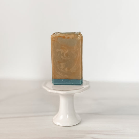 A rectangular bar of Flicker And Twinkle's Olive Branch Goat Milk Soap, marbled in orange and beige with a blue base, sits upright on a small, white pedestal stand against a plain, light-colored background.