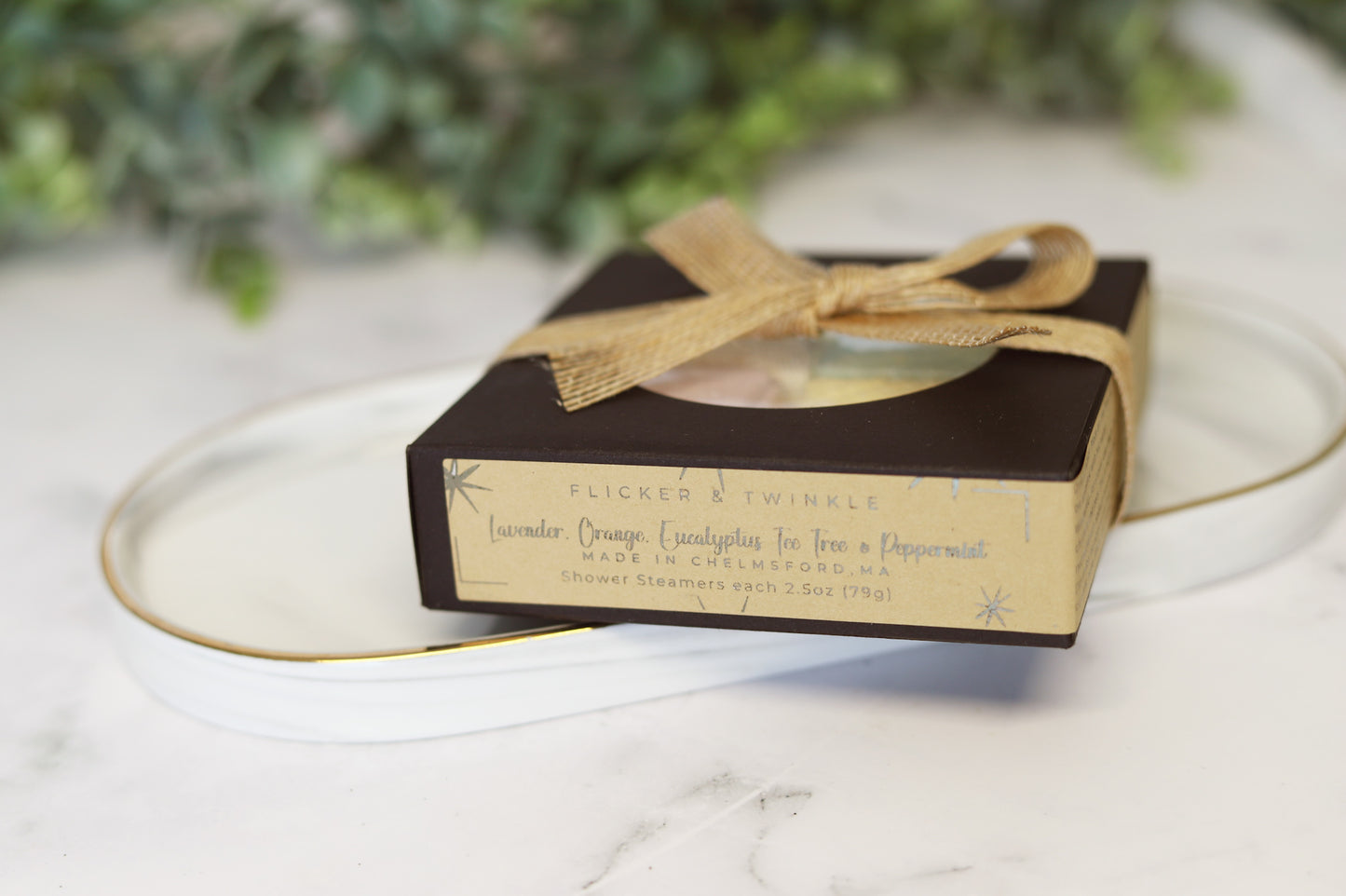 A boxed set of Flicker And Twinkle Shower Steamers with a light brown label, featuring soothing scents such as lavender, orange, eucalyptus, tea tree, and peppermint. The box is elegantly tied with a burlap ribbon and beautifully presented on a white ceramic tray. Blurred greenery in the background adds to the spa-style escape ambiance.