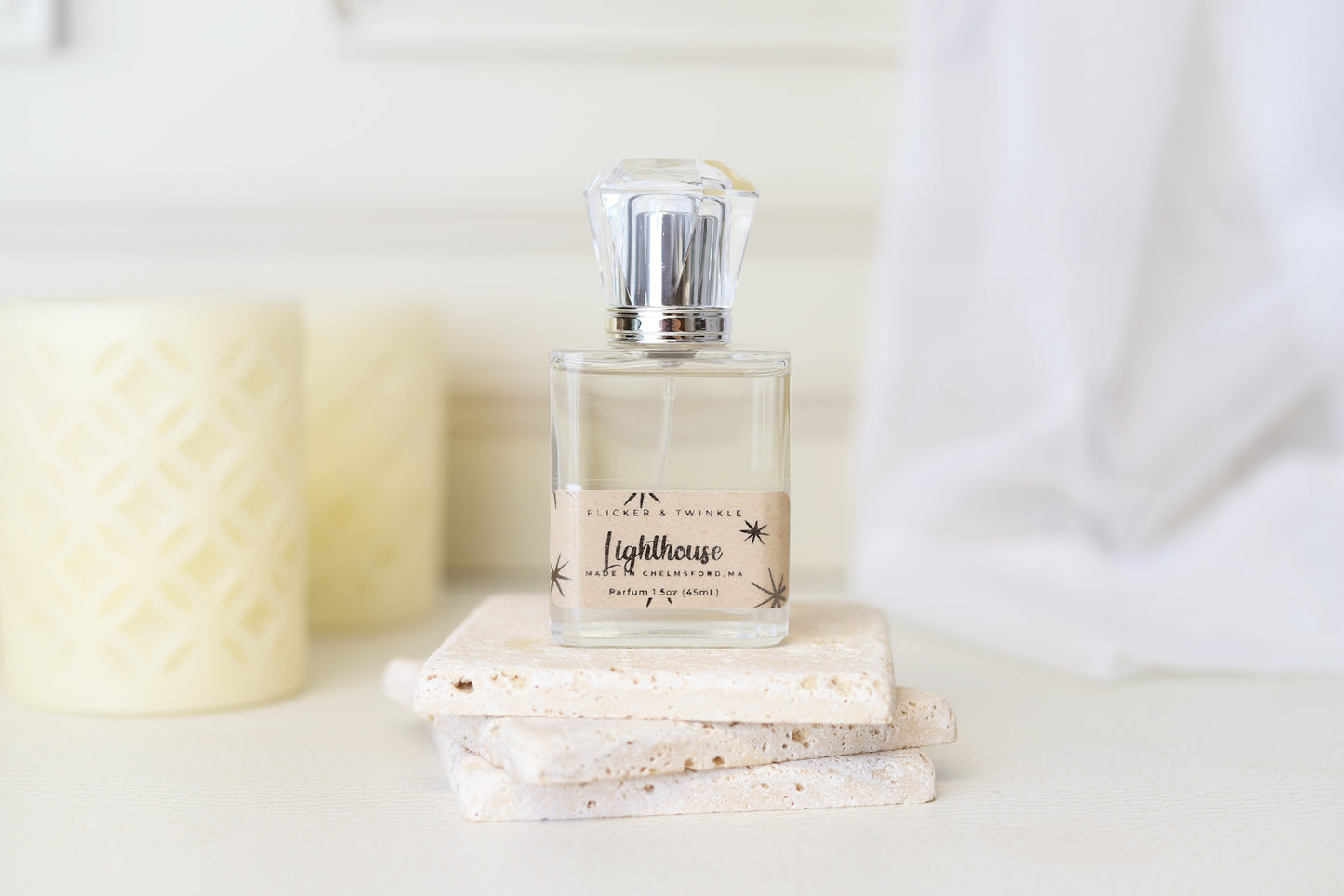 A glass bottle of 'Lighthouse Parfum' by Flicker And Twinkle rests elegantly on two stacked stone tiles, its diamond-shaped cap glinting in the light. Behind it, two light-colored, cylindrical candles adorned with intricate cutouts evoke ocean air, while a white draped fabric completes the serene scene.