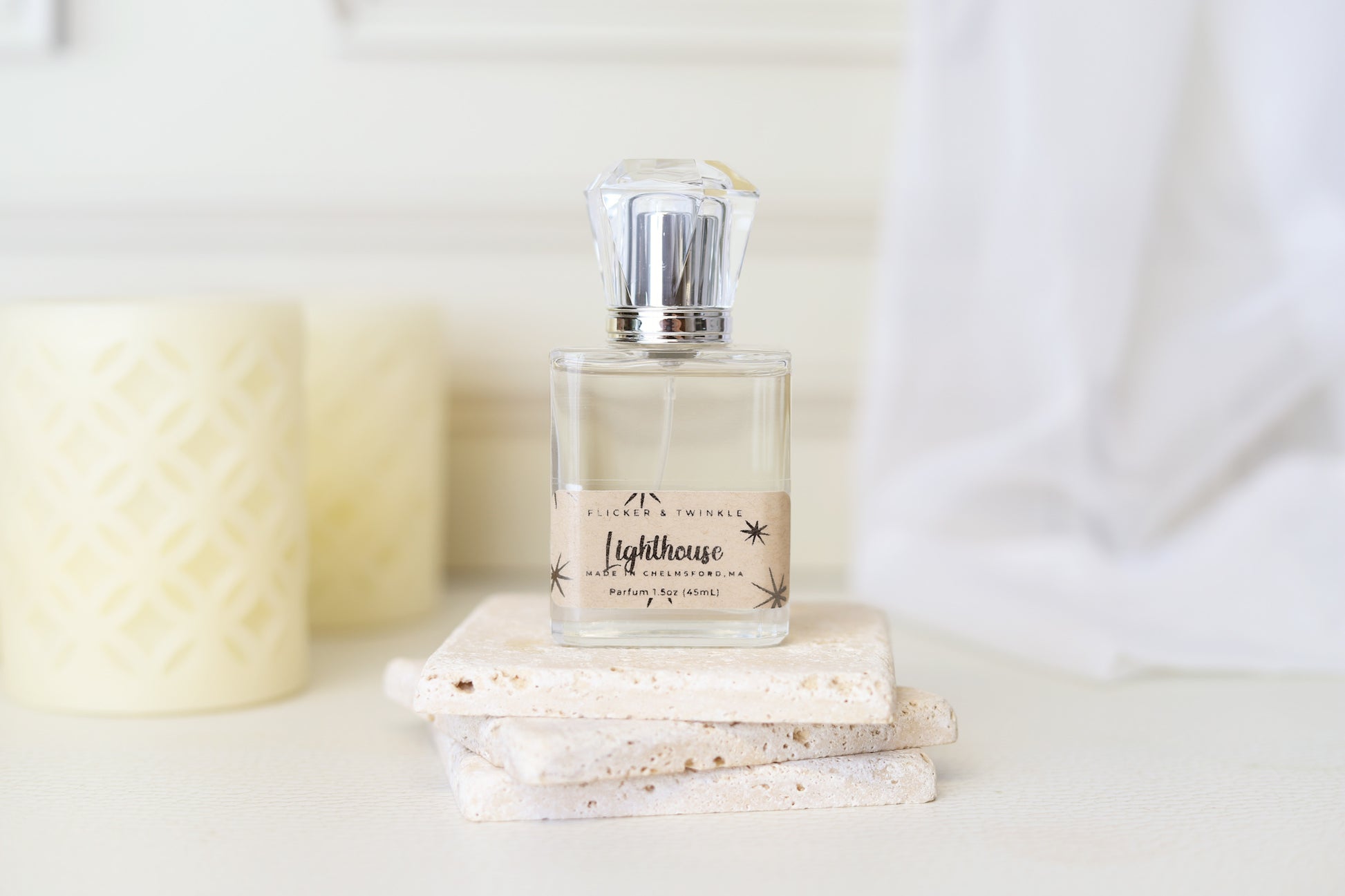 A glass bottle of 'Lighthouse Parfum' by Flicker And Twinkle rests elegantly on two stacked stone tiles, its diamond-shaped cap glinting in the light. Behind it, two light-colored, cylindrical candles adorned with intricate cutouts evoke ocean air, while a white draped fabric completes the serene scene.