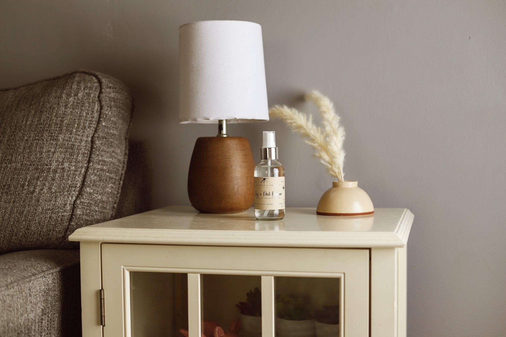 A cozy living room scene featuring a wooden side table with a small brown and white lamp, a bottle of Flicker And Twinkle's Fig and Pink Peppercorn Room and Linen Spray, and a decorative ceramic vase with pampas grass. Next to the table is the arm of a gray upholstered sofa, adding to the luxury home fragrance ambiance.