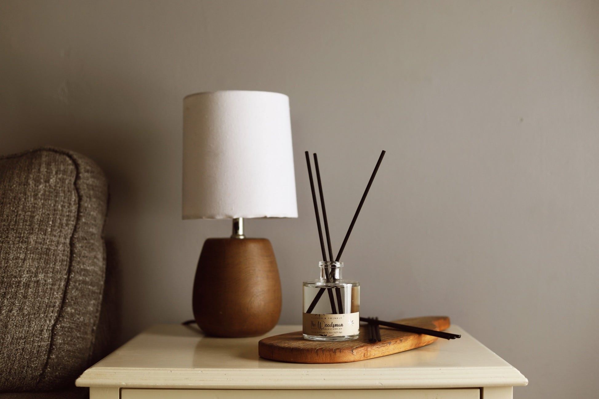 A cozy living room setup featuring a small table lamp with a white shade and wooden base, alongside a wooden tray holding Flicker And Twinkle's The Woodsman Reed Diffuser in a transparent jar with black reeds. The earthy masculine scent complements the beige sofa and neutral wall in the background.